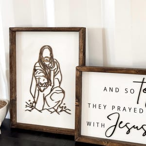 Puede incluir: Dos letreros enmarcados de madera con fondos blancos. El letrero de la izquierda presenta un dise&ntilde;o de corte por l&aacute;ser de Jes&uacute;s sosteniendo a un ni&ntilde;o. El letrero de la derecha presenta texto negro que dice "Y as&iacute; oraron con Jes&uacute;s".