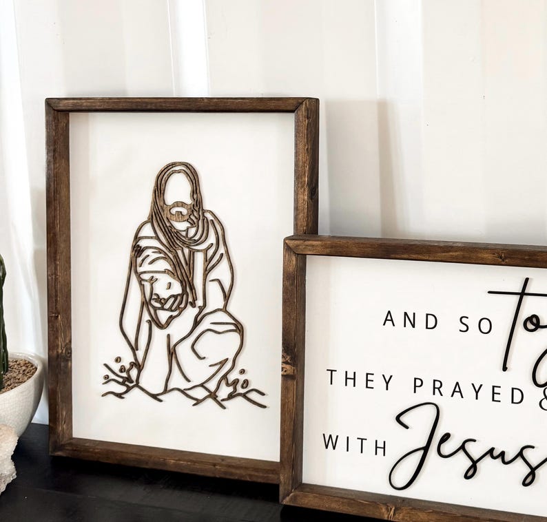 Puede incluir: Dos letreros enmarcados de madera con fondos blancos. El letrero de la izquierda presenta un dise&ntilde;o de corte por l&aacute;ser de Jes&uacute;s sosteniendo a un ni&ntilde;o. El letrero de la derecha presenta texto negro que dice "Y as&iacute; oraron con Jes&uacute;s".
