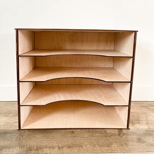 Könnte beinhalten: Ein Holzorganizer mit vier Regalen. Die Regale haben eine gebogene Vorderkante und sind aus hellem Holz gefertigt. Der Organizer steht auf einem Holzboden.