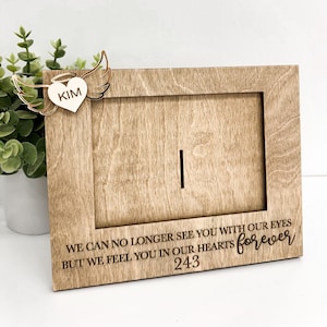 May include: Wooden picture frame with a heart-shaped cutout and angel wings. The heart has the name "KIM" engraved on it. The frame has the text "WE CAN NO LONGER SEE YOU WITH OUR EYES BUT WE FEEL YOU IN OUR HEARTS forever 243" engraved on the bottom.