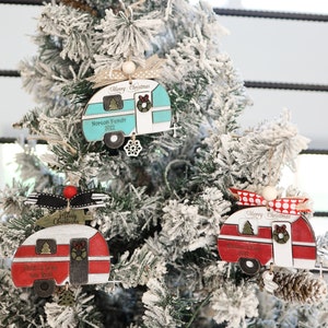 May include: Three Christmas ornaments shaped like vintage campers. The ornaments are made of wood and painted in red, blue, and white. Each ornament has a Merry Christmas message and a personalized family name. The ornaments are hanging on a snowy Christmas tree.