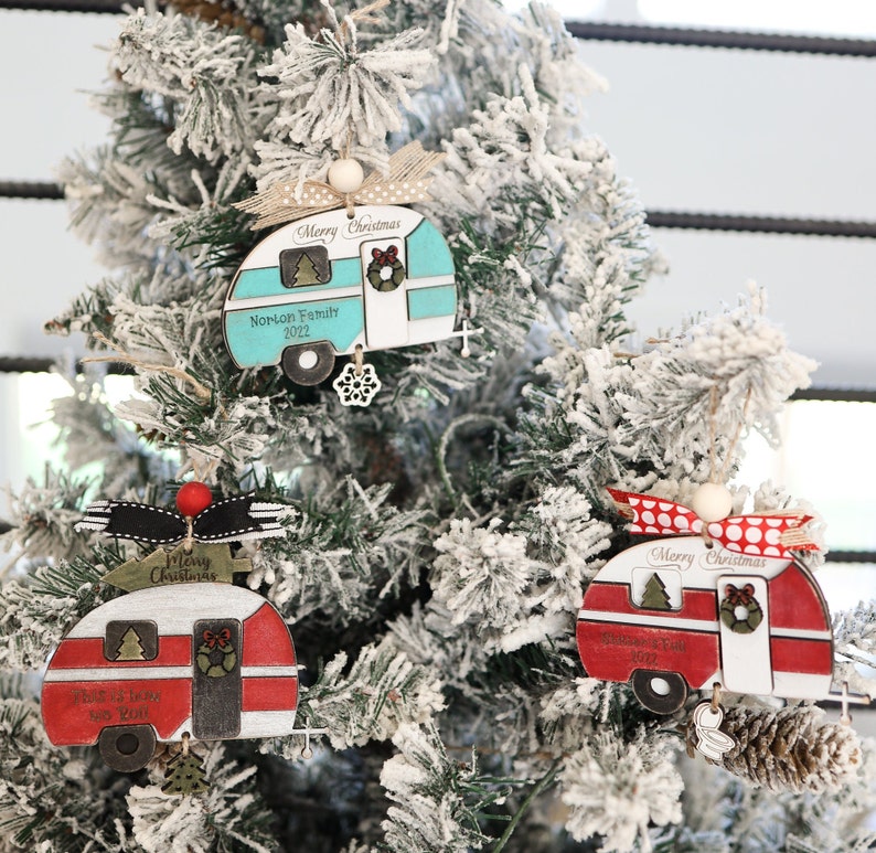May include: Three Christmas ornaments shaped like vintage campers. The ornaments are made of wood and painted in red, blue, and white. Each ornament has a Merry Christmas message and a personalized family name. The ornaments are hanging on a snowy Christmas tree.