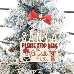 May include: Wooden Christmas ornament with the text "Santa Please Stop Here For Charlotte & Ella". The ornament features a red bow, a Santa Claus figure, and a reindeer.