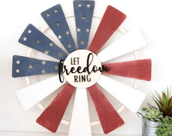 Può includere: Un mulino a vento in legno a tema bandiera americana con vernice rossa, bianca e blu. Il mulino a vento ha una base circolare e 12 pale. Le pale sono dipinte in colori alternati di rosso, bianco e blu. Il centro del mulino a vento ha un cartello che recita "Let Freedom Ring".