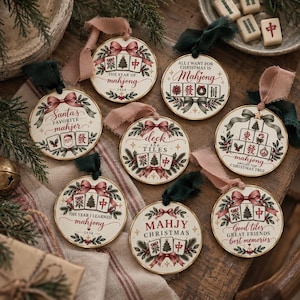 May include: Eight round ceramic ornaments featuring festive illustrations of mahjong tiles, Christmas trees, and holly. Each ornament displays unique holiday-themed text such as The Year of Mahjong 2026, Santa's Favorite Mahjer, and Deck the Tiles, finished with ribbons.