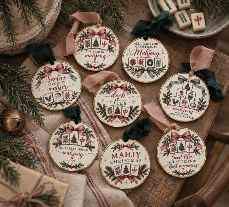 May include: Eight round ceramic ornaments featuring festive illustrations of mahjong tiles, Christmas trees, and holly. Each ornament displays unique holiday-themed text such as The Year of Mahjong 2026, Santa's Favorite Mahjer, and Deck the Tiles, finished with ribbons.