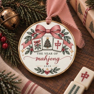 May include: A round ceramic ornament with a gold rim and a pink ribbon, featuring three mahjong tiles, a pink bow, holly leaves, and berries. The text reads THE YEAR OF mahjong 2026, decorated with small stars and a heart icon.