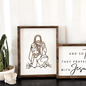 Puede incluir: Dos letreros de madera con fondo blanco y marcos marrones. El letrero de la izquierda presenta un dise&ntilde;o de corte por l&aacute;ser de Jes&uacute;s llevando a una persona en la espalda. El letrero de la derecha tiene el texto "And so together they prayed & built with Jesus at".
