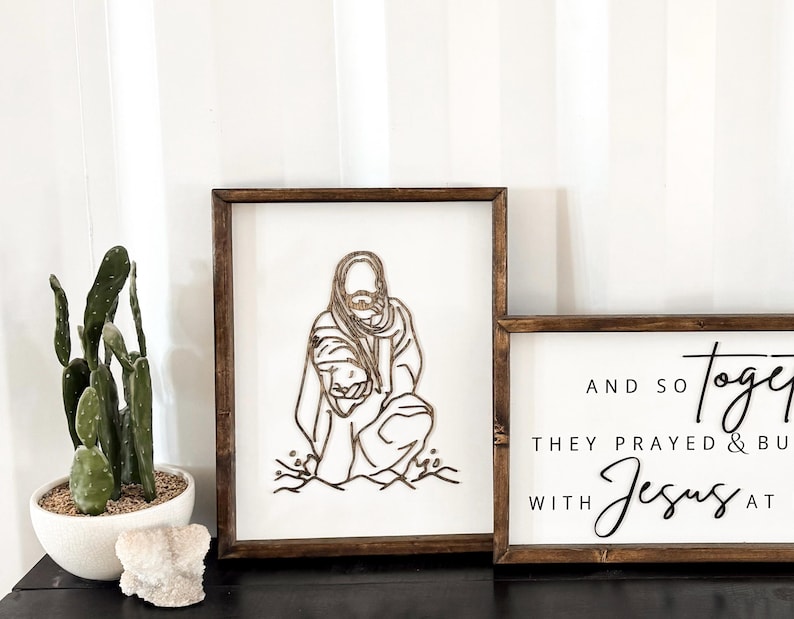 Puede incluir: Dos letreros de madera con fondo blanco y marcos marrones. El letrero de la izquierda presenta un dise&ntilde;o de corte por l&aacute;ser de Jes&uacute;s llevando a una persona en la espalda. El letrero de la derecha tiene el texto "And so together they prayed & built with Jesus at".