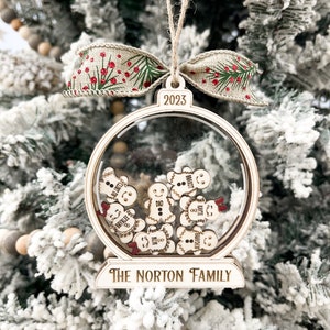 May include: A wooden Christmas bauble shaped like a snow globe with a clear plastic dome. The dome contains small wooden gingerbread people with names written on them. The bauble is decorated with the year 2023 and the text "The Norton Family".