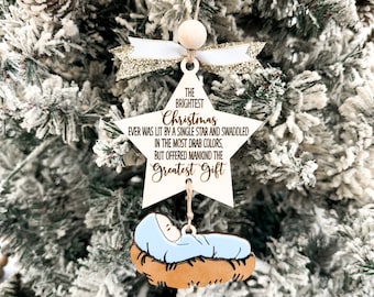 May include: A wooden star ornament with a white background and gold glitter accents. The star features the text "The Brightest Christmas Ever Was Lit By A Single Star And Swaddled In The Most Drab Colors, But Offered Mankind The Greatest Gift". A small wooden baby Jesus figure is hanging from the star.