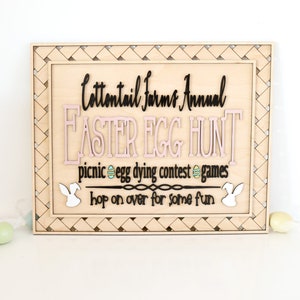 Puede incluir: Un letrero de madera con un borde tejido, anunciando una b&uacute;squeda anual de huevos de Pascua de Cottontail Farms. El letrero presenta letras rosas y negras, y el texto dice: "Cottontail Farms Annual Easter Egg Hunt, picnic egg dying contest games, hop on over for some fun."