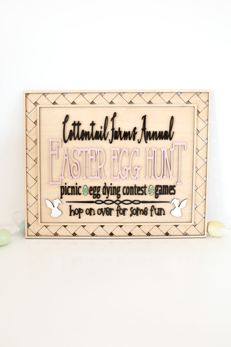 Puede incluir: Un letrero de madera con un borde tejido, anunciando una b&uacute;squeda anual de huevos de Pascua de Cottontail Farms. El letrero presenta letras rosas y negras, y el texto dice: "Cottontail Farms Annual Easter Egg Hunt, picnic egg dying contest games, hop on over for some fun."