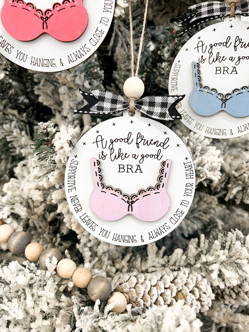 May include: A white round ornament with a pink bra cutout and the text "A good friend is like a good BRA" with the text "SUPPORTIVE. NEVER LEAVES YOU HANGING, & ALWAYS CLOSE TO YOUR HEART" around the edge.