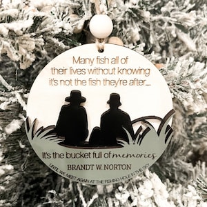May include: A wooden Christmas ornament with a silhouette of two people fishing. The ornament has the text "Many fish all of their lives without knowing it's not the fish they're after... It's the bucket full of memories BRANDT W. NORTON UNTIL WE MEET AGAIN AT THE FISHING HOLE IN THE SKY".
