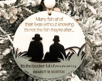 May include: A wooden Christmas ornament with a silhouette of two people fishing. The ornament has the text "Many fish all of their lives without knowing it's not the fish they're after... It's the bucket full of memories BRANDT W. NORTON UNTIL WE MEET AGAIN AT THE FISHING HOLE IN THE SKY".