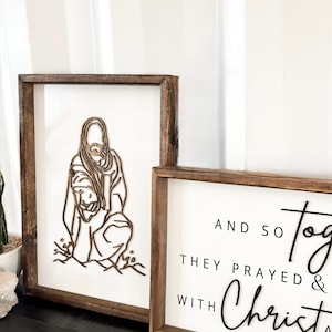 Puede incluir: Dos letreros enmarcados en madera con fondos blancos. El letrero de la izquierda presenta un dise&ntilde;o de madera cortado con l&aacute;ser de Jes&uacute;s arrodillado con un ni&ntilde;o. El letrero de la derecha tiene texto negro que dice "And so they prayed & with Christ".