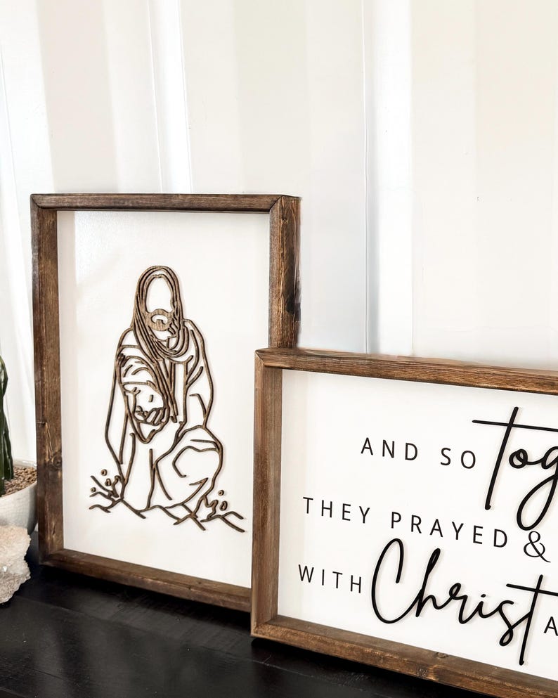 Puede incluir: Dos letreros enmarcados en madera con fondos blancos. El letrero de la izquierda presenta un dise&ntilde;o de madera cortado con l&aacute;ser de Jes&uacute;s arrodillado con un ni&ntilde;o. El letrero de la derecha tiene texto negro que dice "And so they prayed & with Christ".