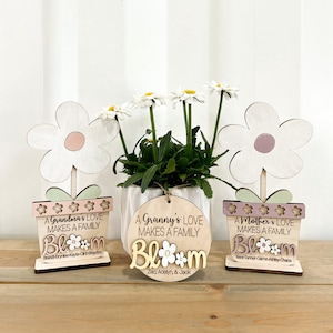 May include: Three wooden flower pot decorations with the text "A Grandma's Love Makes a Family", "A Granny's Love Makes a Family", and "A Mother's Love Makes a Family". Each pot has a white flower with a pink, purple, or peach centre. The text "Bloom" is written below each pot with the names of family members.