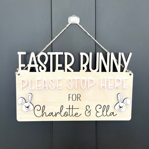 Könnte beinhalten: Ein Holzschild mit dem Text "Easter Bunny Please Stop Here For Charlotte & Ella" mit zwei Hasenschnitten auf beiden Seiten der Namen. Das Schild hängt an einer Schnur.