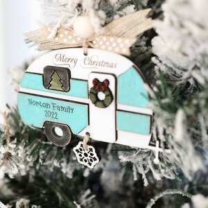 May include: A teal and white wooden camper Christmas ornament with the text "Merry Christmas" and "Norton Family 2022". The ornament has a small green tree, a red bow, and a green wreath on the front. A snowflake charm hangs from the bottom.