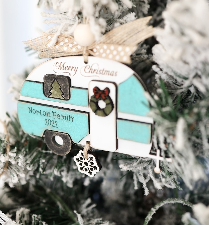May include: A teal and white wooden camper Christmas ornament with the text "Merry Christmas" and "Norton Family 2022". The ornament has a small green tree, a red bow, and a green wreath on the front. A snowflake charm hangs from the bottom.