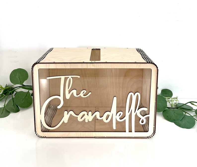 May include: A wooden box with a clear acrylic front and a slot on the top. The box has the words "The Crandells" written on the front in white.