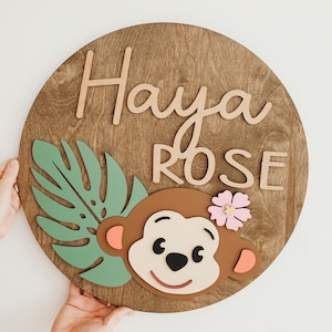 May include: A round wooden sign with the name "Haya Rose" written in cursive script. A green leaf and a brown monkey with a pink flower are attached to the sign.