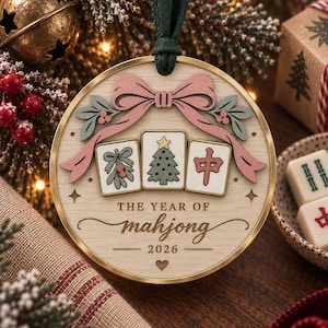May include: A round wooden Christmas ornament with a gold metal rim and a dark green ribbon hanger. The ornament features a pink bow, three mahjong tiles with holiday designs, and the text "THE YEAR OF mahjong 2026" with a small heart icon.