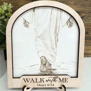 May include: A wooden framed art piece with a pencil sketch of a figure walking. The light-coloured wooden frame has the words "WALK with ME" and "Moses 6:34" etched on the bottom. The art is displayed on a small wooden stand.