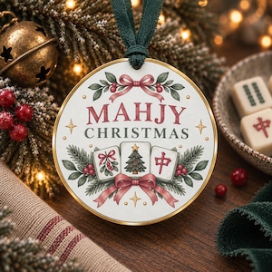 May include: A round white Christmas ornament with a gold rim and a dark green ribbon hanger. The design features red bows, green pine branches, and three mahjong tiles, with the text "MAHJY CHRISTMAS" printed in red and dark green lettering.