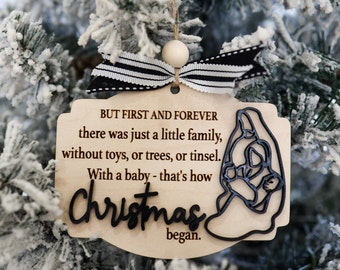 May include: A wooden Christmas ornament with a black and white striped ribbon. The ornament features a silhouette of Mary holding baby Jesus with the text "BUT FIRST AND FOREVER there was just a little family, without toys, or trees, or tinsel. With a baby - that's how Christmas began."
