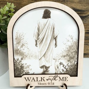 May include: A wooden framed art piece featuring a sepia-toned illustration of a figure walking with a lamb. The text "WALK with ME" and "Moses 6:34" are printed at the bottom. The frame has a built-in stand.