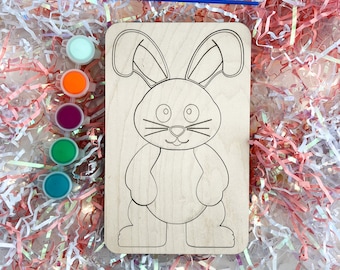 May include: A wooden craft kit featuring a bunny outline ready to be painted. The kit includes four small paint pots in various colors: light blue, orange, purple, and green.