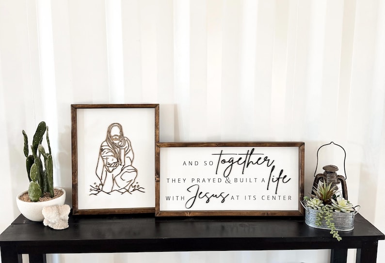 Puede incluir: Dos letreros de madera con fondo blanco y texto negro. El primer letrero presenta un dibujo lineal de una persona con un ni&ntilde;o. El segundo letrero dice "AND SO TOGETHER THEY PRAYED & BUILT A LIFE WITH JESUS AT ITS CENTER".