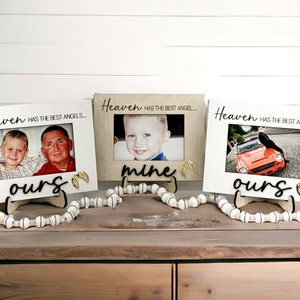 May include: Three white wooden picture frames with the text "Heaven has the best angels" and "Heaven has the best angel" on each frame. The frames are decorated with gold angel wings and the words "ours", "mine", and "ours" are written below the photos. The frames are sitting on a wooden surface with white beads.