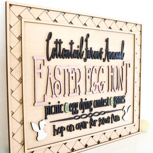 Puede incluir: Un letrero de madera con un borde tejido, anunciando una "Cottontail Farms Annual Easter Egg Hunt". El letrero presenta un texto "Easter Egg Hunt" rosa y negro, con un texto m&aacute;s peque&ntilde;o debajo que dice "picnic egg dying contest games hop on over for some fun".