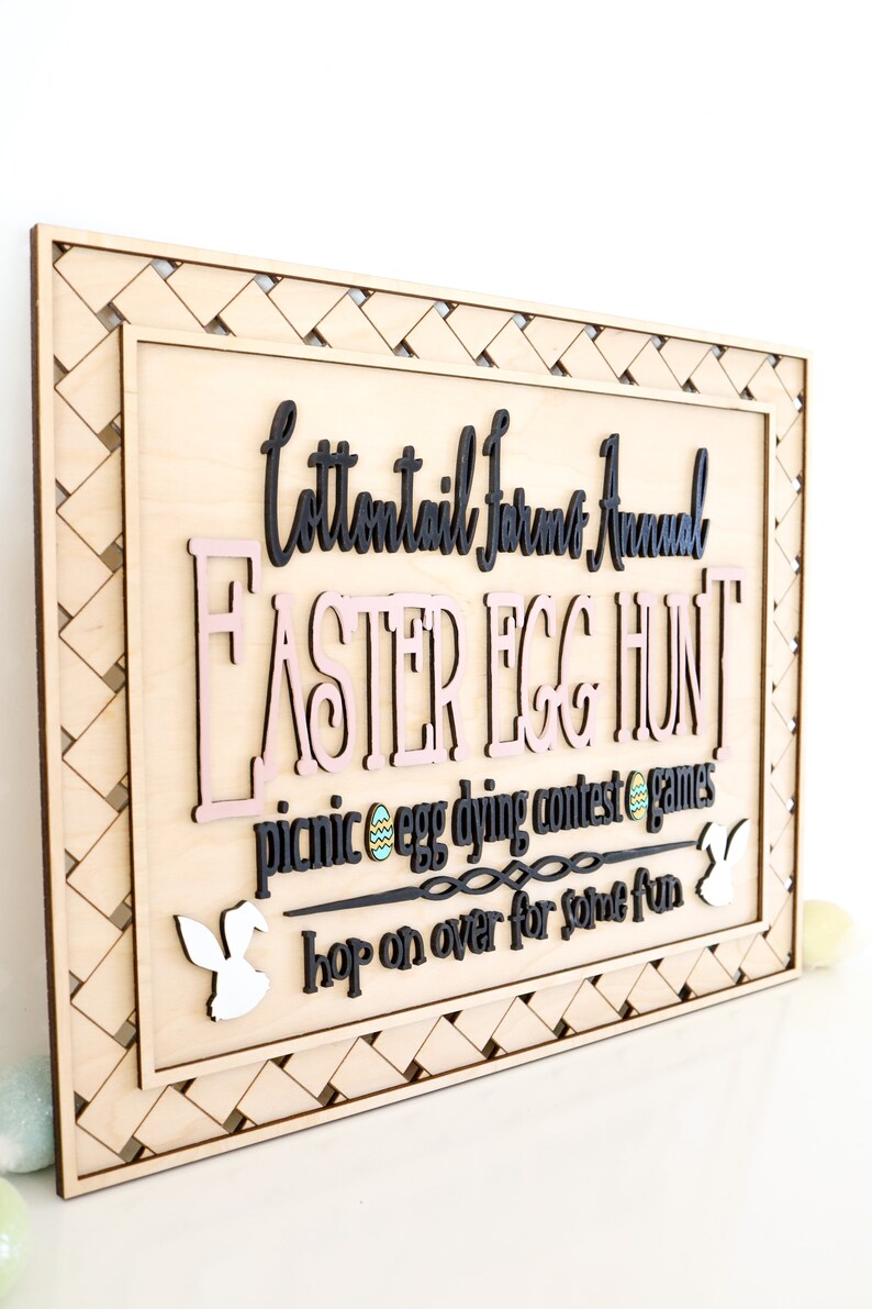 Puede incluir: Un letrero de madera con un borde tejido, anunciando una "Cottontail Farms Annual Easter Egg Hunt". El letrero presenta un texto "Easter Egg Hunt" rosa y negro, con un texto m&aacute;s peque&ntilde;o debajo que dice "picnic egg dying contest games hop on over for some fun".