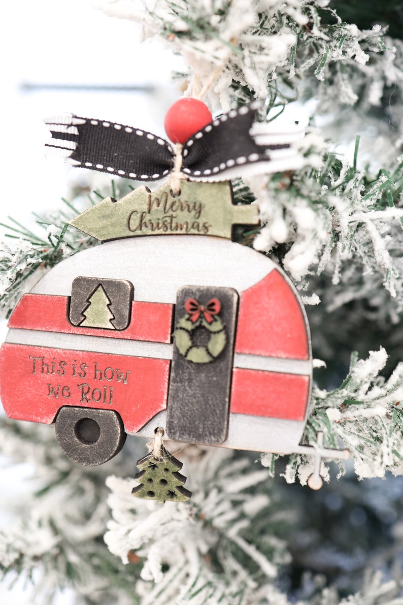 May include: A wooden Christmas ornament shaped like a camper with the text "Merry Christmas" and "This is how we roll". The camper is red, white, and black with a green Christmas tree on top.