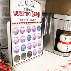 May include: A wooden sign with the text "hot chocolate is like a warm hug from the inside" and a row of coffee pods in front of it. The coffee pods are in a holder with a white background and are arranged in a grid pattern. The sign is on a white countertop with a coffee maker and a snowman mug in the background.