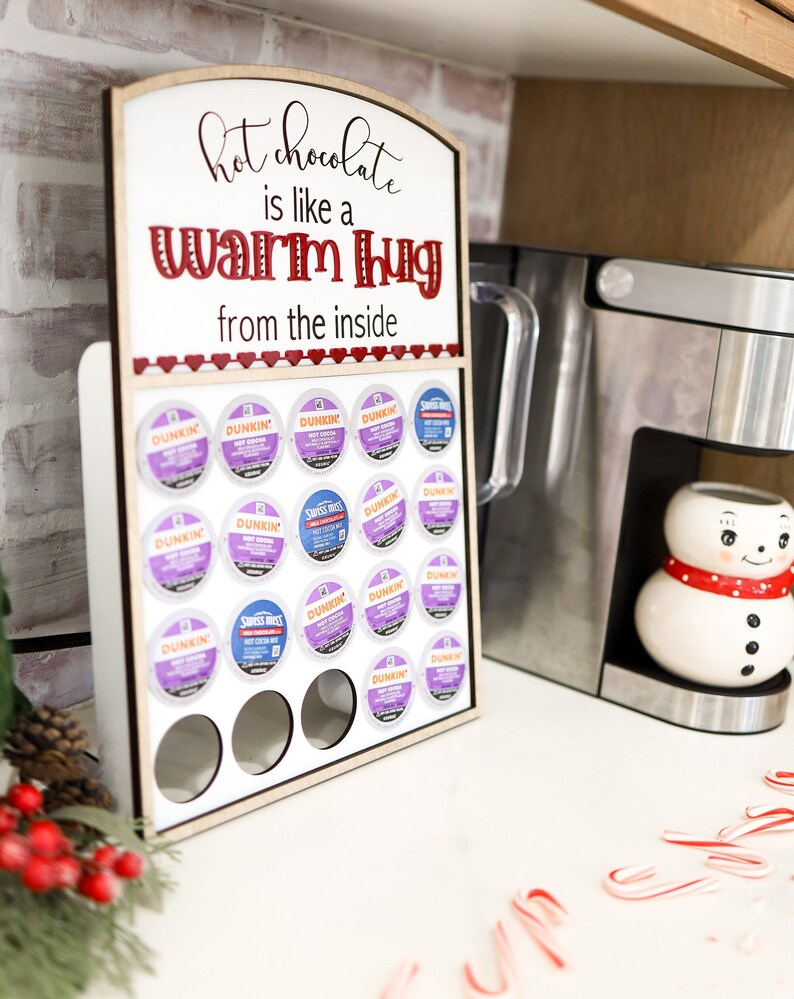 May include: A wooden sign with the text "hot chocolate is like a warm hug from the inside" and a row of coffee pods in front of it. The coffee pods are in a holder with a white background and are arranged in a grid pattern. The sign is on a white countertop with a coffee maker and a snowman mug in the background.