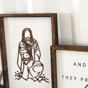 Puede incluir: Dos letreros enmarcados de madera con fondo blanco. El letrero de la izquierda presenta un dise&ntilde;o de corte por l&aacute;ser de Jes&uacute;s sosteniendo a un ni&ntilde;o. El letrero de la derecha tiene el texto "Y ORARON CON DIOS".