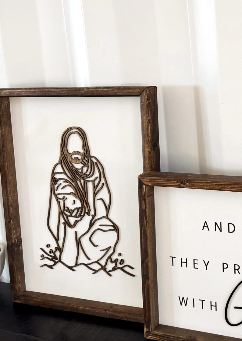 Puede incluir: Dos letreros enmarcados de madera con fondo blanco. El letrero de la izquierda presenta un dise&ntilde;o de corte por l&aacute;ser de Jes&uacute;s sosteniendo a un ni&ntilde;o. El letrero de la derecha tiene el texto "Y ORARON CON DIOS".