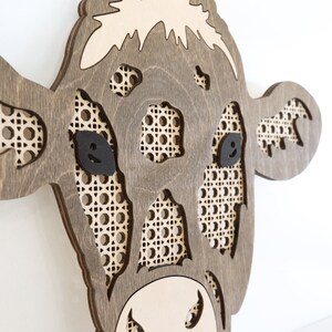 K&ouml;nnte beinhalten: Eine Wanddekoration aus Holz in Form eines Kuhkopfes mit brauner Beize. Die Augen der Kuh sind schwarz und Nase und Maul sind aus dem Holz ausgeschnitten. Das Gesicht der Kuh ist mit einem geflochtenen Rohrgeflecht verziert.