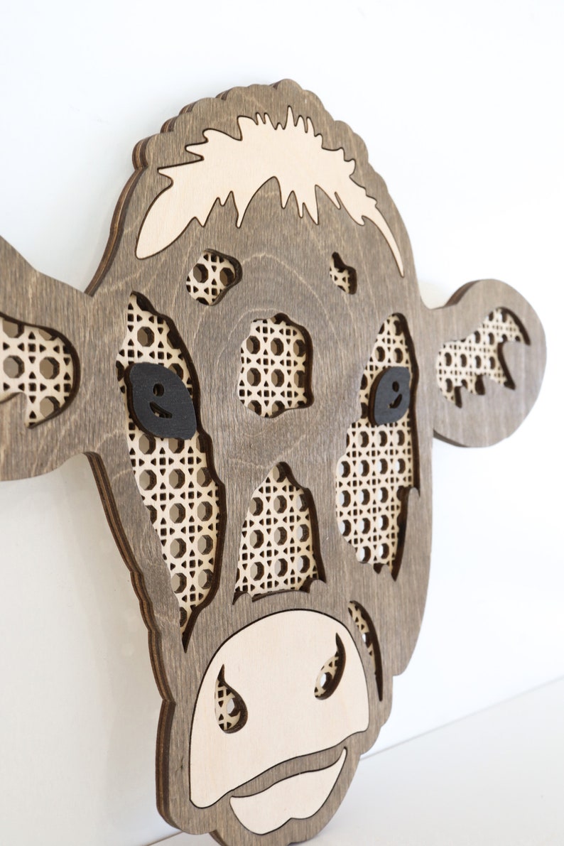 K&ouml;nnte beinhalten: Eine Wanddekoration aus Holz in Form eines Kuhkopfes mit brauner Beize. Die Augen der Kuh sind schwarz und Nase und Maul sind aus dem Holz ausgeschnitten. Das Gesicht der Kuh ist mit einem geflochtenen Rohrgeflecht verziert.