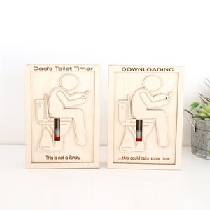 Puede incluir: Dos letreros de madera con la silueta de una persona sentada en el inodoro. El letrero de la izquierda dice "Dad's Toilet Timer" y "This is not a library". El letrero de la derecha dice "DOWNLOADING" y "...this could take some time".