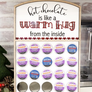 May include: A wooden stand with a sign that reads "hot chocolate is like a warm hug from the inside". The stand holds 18 single-serve coffee pods, including Dunkin' Donuts and Swiss Miss hot cocoa.