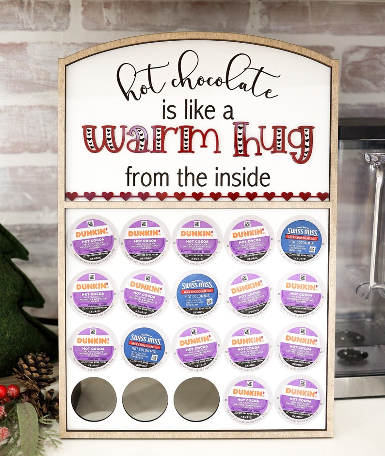 May include: A wooden stand with a sign that reads "hot chocolate is like a warm hug from the inside". The stand holds 18 single-serve coffee pods, including Dunkin' Donuts and Swiss Miss hot cocoa.