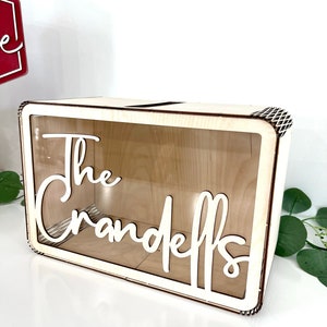 May include: A wooden box with a clear acrylic front and a slot on the top. The box has the words "The Crandells" cut out in white lettering.