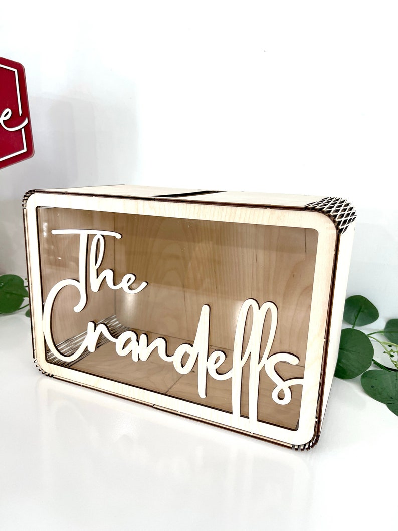 May include: A wooden box with a clear acrylic front and a slot on the top. The box has the words "The Crandells" cut out in white lettering.